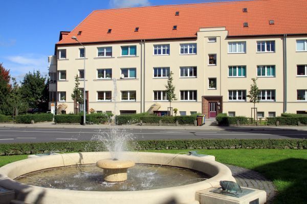 Helle Wohnhausfassade mit rotem Satteldach, im Vordergrund ein runder Brunnen mit sprudelndem Wasserstrahl, umgeben von gepflegten Grünflächen. Die Szene wirkt freundlich und einladend bei blauem Himmel und Sonnenschein.