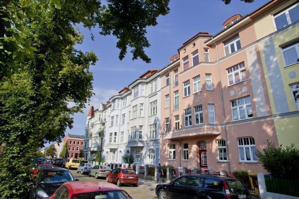 Reihe farbenfroher Altbauten mit reich verzierten Fassaden und roten Dächern, die im Sonnenlicht strahlen. Entlang der ruhigen Straße stehen parkende Autos, während Bäume im Vordergrund Schatten spenden und der Szene eine lebendige, zugleich gemütliche Atmosphäre verleihen.