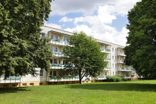 Heller Wohnblock mit grünen Balkonbrüstungen, teils geschmückt mit Blumenkästen. Vor dem Gebäude breitet sich eine großzügige Rasenfläche mit hohen, schattenspendenden Bäumen aus, die der Szene eine ruhige, naturnahe Atmosphäre verleihen.