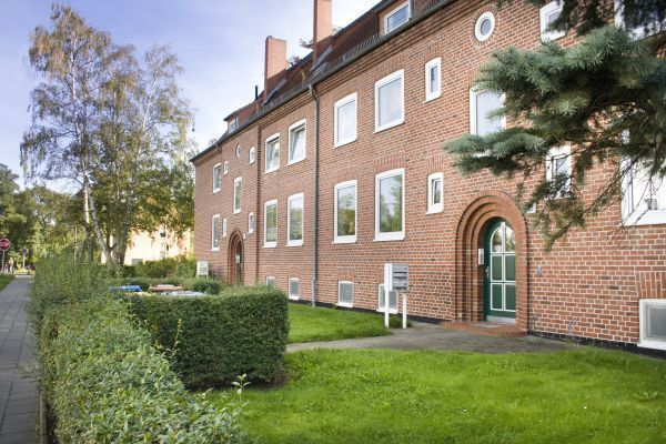 Backsteinwohnhaus mit grüner Eingangstür und dekorativen Rundbögen, umgeben von gepflegten Hecken und Rasenflächen. Der Gehweg und die Bäume am Straßenrand verleihen der Szene einen ruhigen, wohnlichen Charakter.