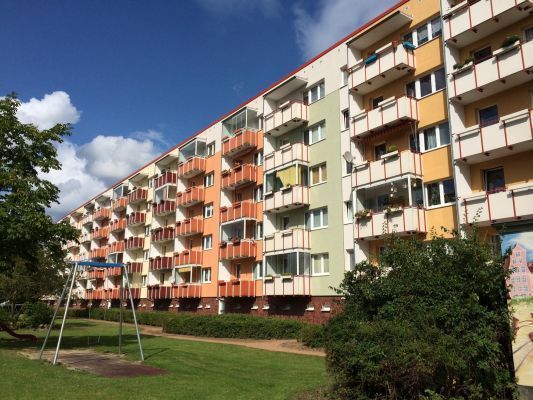 Farbenfroher Wohnblock mit Balkonen in warmen Orange- und Gelbtönen, davor eine gepflegte Rasenfläche mit Spielgeräten. Der blaue Himmel mit weißen Wolken unterstreicht die freundliche, wohnliche Atmosphäre.