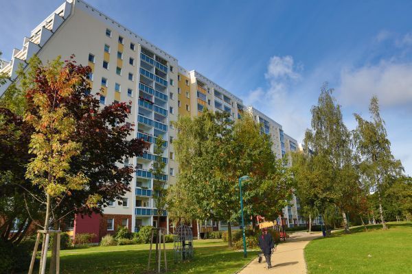 Mehrstöckiges Wohnhaus mit hellen Fassaden und bunten Balkonfronten, umgeben von einer grünen Parkanlage mit Bäumen und Wiese. Ein gepflasterter Weg führt durch die Anlage, auf dem Menschen spazieren gehen, während herbstliche Sonnenstrahlen die Szene warm erleuchten.