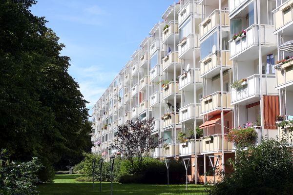 Langer Wohnblock mit weißen Balkonen, von denen viele bepflanzt oder mit Markisen geschmückt sind. Im Vordergrund liegt eine weitläufige Grünfläche mit Bäumen und Sträuchern, die der Szene eine ruhige und wohnliche Atmosphäre verleihen.