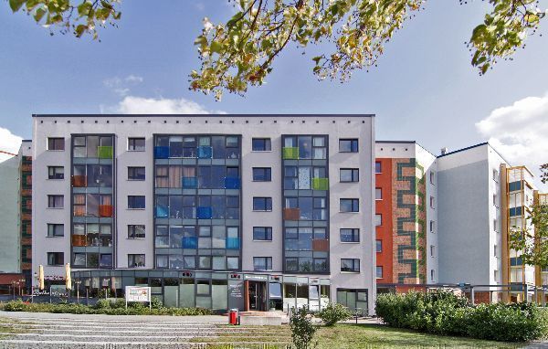 Modernisierter Wohnblock mit bunter Glasfassade und klaren Linien, eingerahmt von Bäumen im Vordergrund. Vor dem Gebäude liegen ein kleiner Platz und Grünflächen, während sich hinter den Fenstern leuchtende Farbflächen abzeichnen, die dem Bauwerk eine lebendige Ausstrahlung geben.