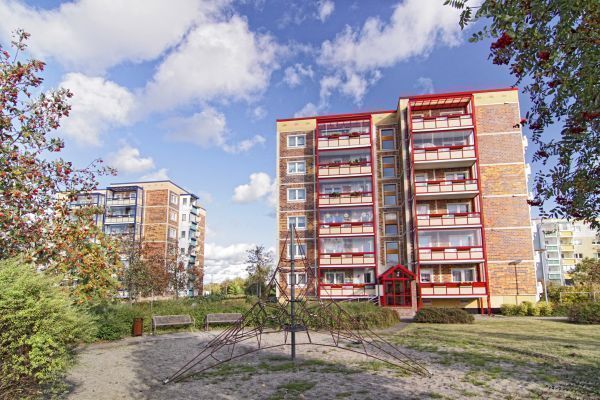 Mehrgeschossiges Wohnhaus mit roten Balkonelementen, davor ein kleiner Platz mit Klettergerüst, Bänken und Grünflächen. Bäume mit herbstlich gefärbtem Laub rahmen die Szene, während blauer Himmel und weiße Wolken eine freundliche Atmosphäre schaffen.