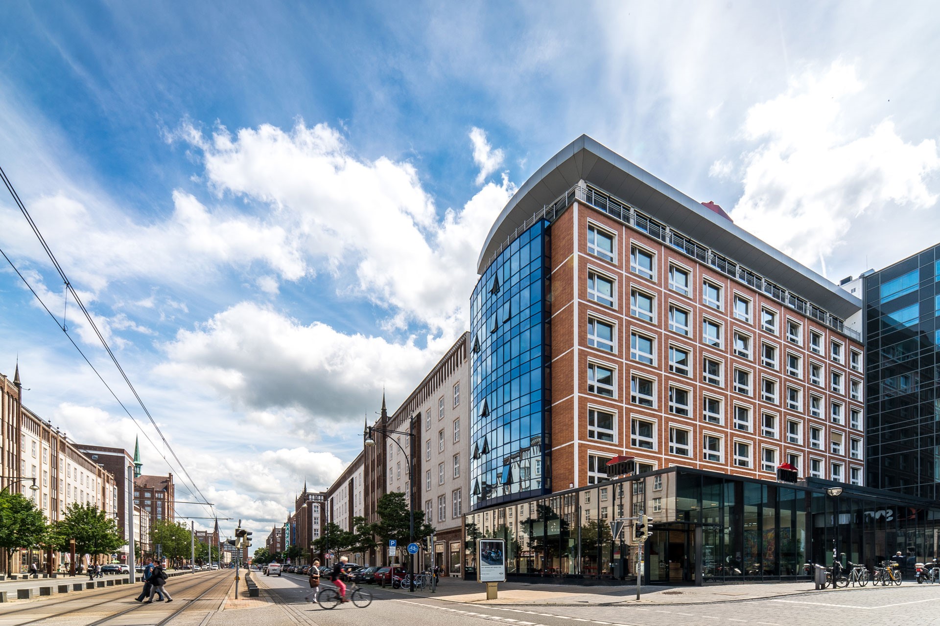 Belebte Straßenszene mit modernen und historischen Gebäuden im Kontrast: Im Vordergrund ein Eckgebäude mit roter Backsteinfassade und markanter Glasfront, dahinter eine Reihe klassischer Fassaden mit Türmen und Ornamenten. Straßenbahntrassen, Radfahrer und Fußgänger beleben die Szenerie unter einem weiten Himmel mit hellen Wolken.