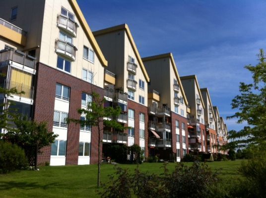 Reihe moderner Wohnhäuser mit markanten Satteldächern und Balkonen, im Vordergrund eine weite, gepflegte Rasenfläche mit jungen Bäumen und Sträuchern. Der blaue Himmel und das Sonnenlicht verleihen der Szene eine offene und freundliche Atmosphäre.