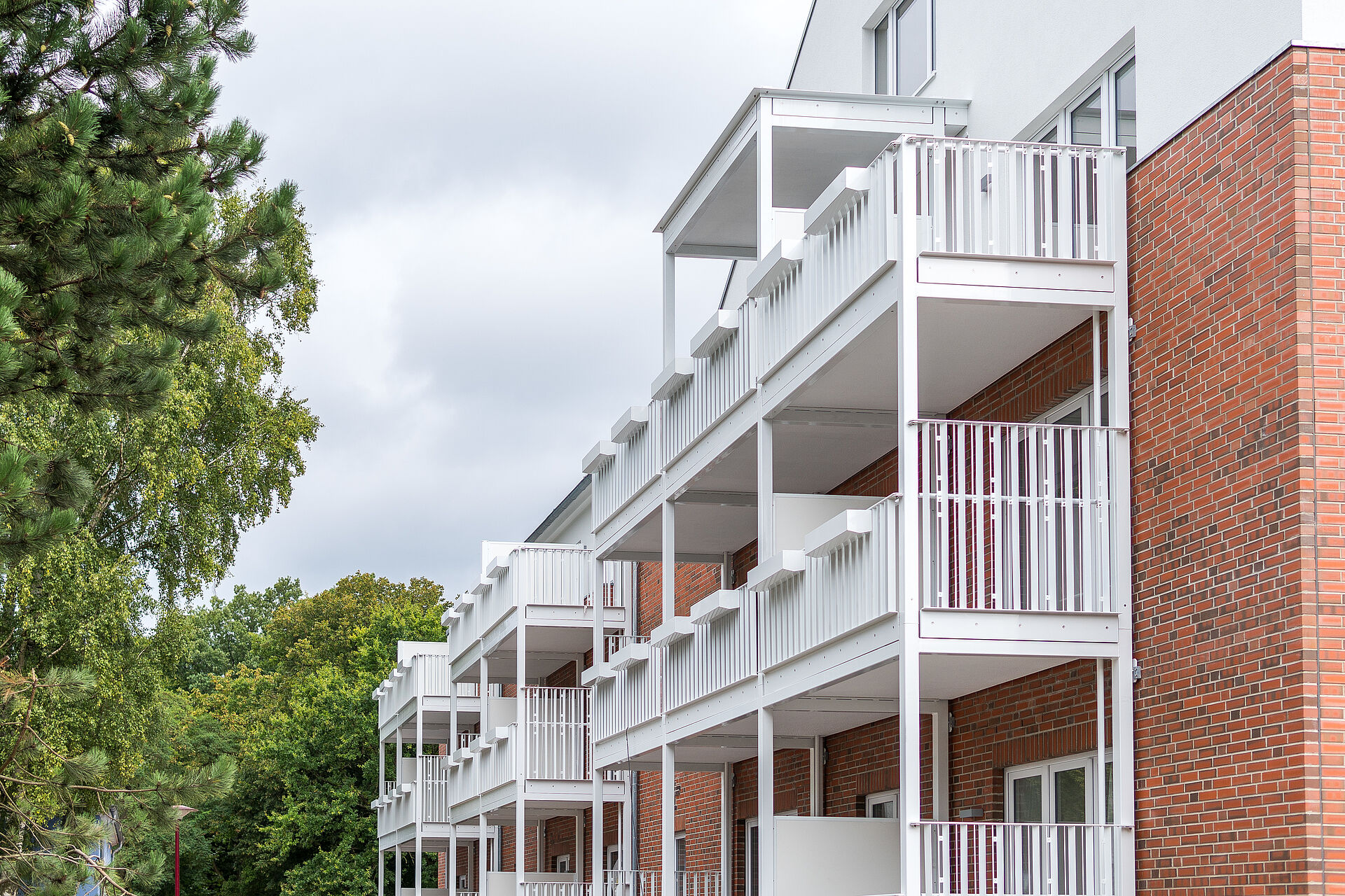 Neubau mit hellen, weißen Balkonen vor einer Fassade aus rotem Klinker, eingerahmt von Bäumen und grünem Laub. Die klare Architektur und das Zusammenspiel von Holzoptik und Weiß erzeugen eine moderne, freundliche Anmutung.