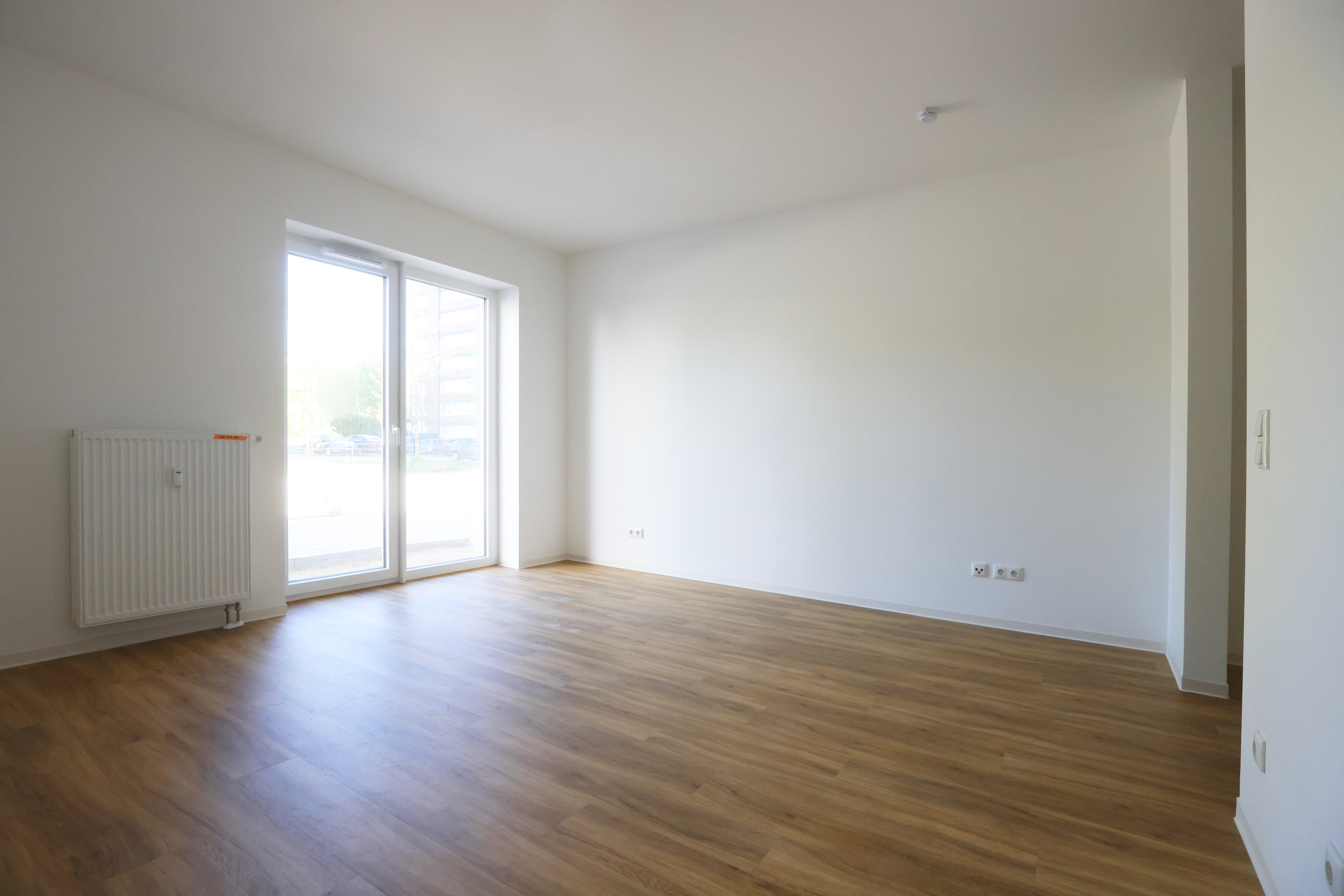 Helles Zimmer mit Fußboden in Holzoptik, breitem, bodentiefem Doppelfenster, Heizkörper und viel freier Stellfläche entlang der Wände.