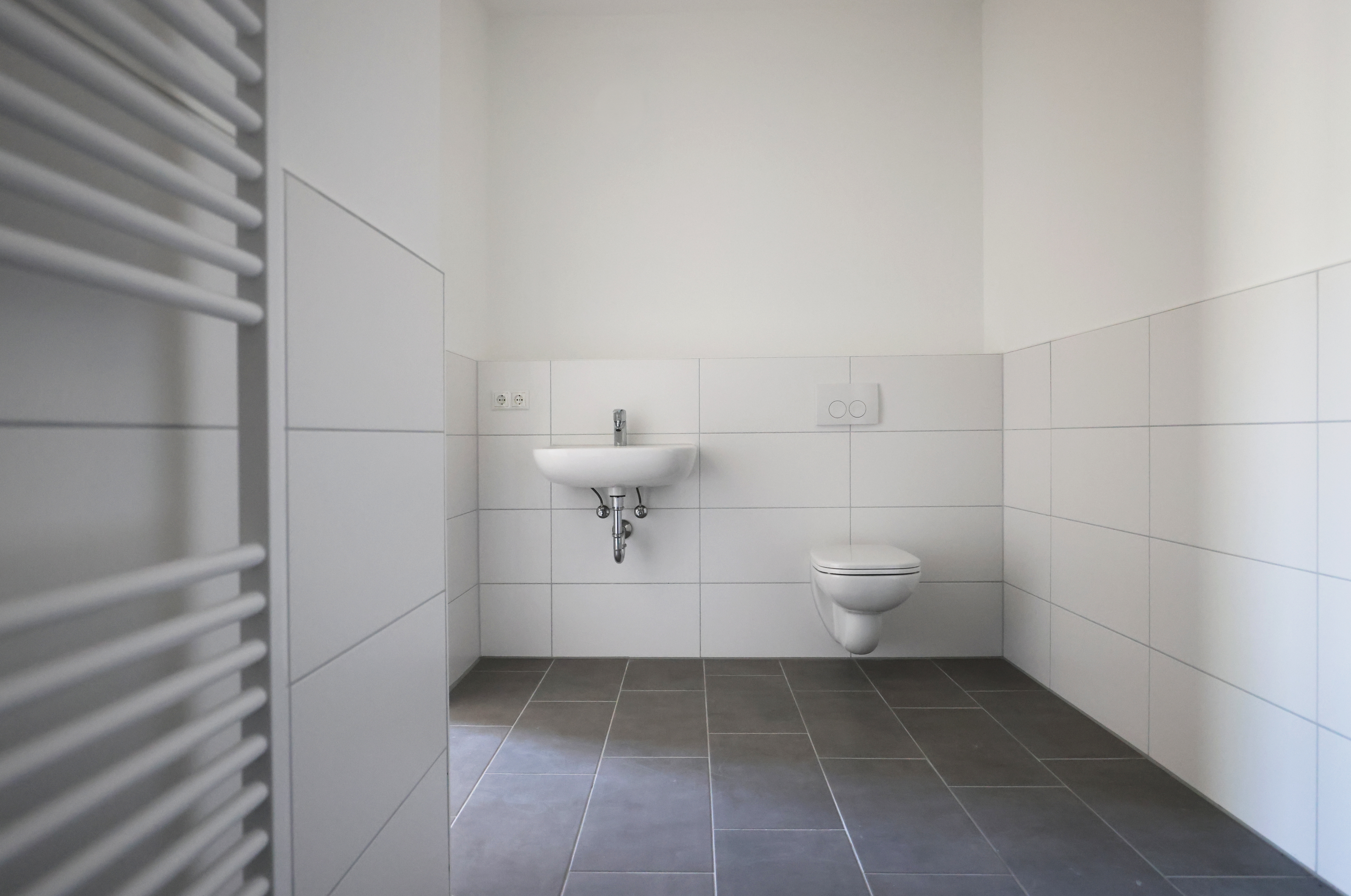 Helles Badezimmer mit Waschbecken, wandhängendem WC, grauem Fliesenboden, weißen Wandfliesen und viel Bewegungsfläche.
