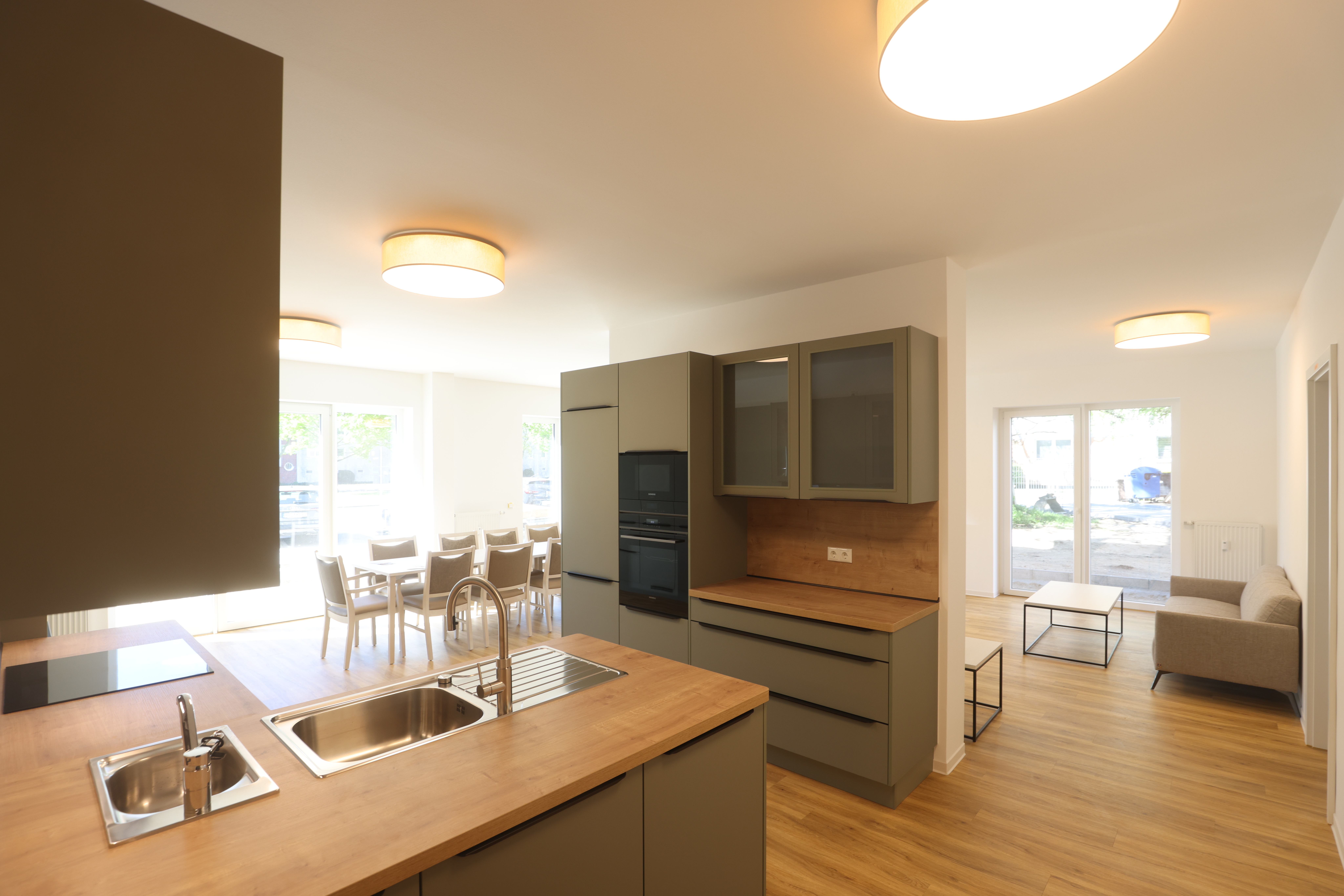 Gemeinschaftsbereich der zweiten betreuten Wohngemeinschaft mit grün-grauer offener Küche, Arbeitsplatten in Holzoptik, großem Esstisch und Wohnbereich mit Sofa.