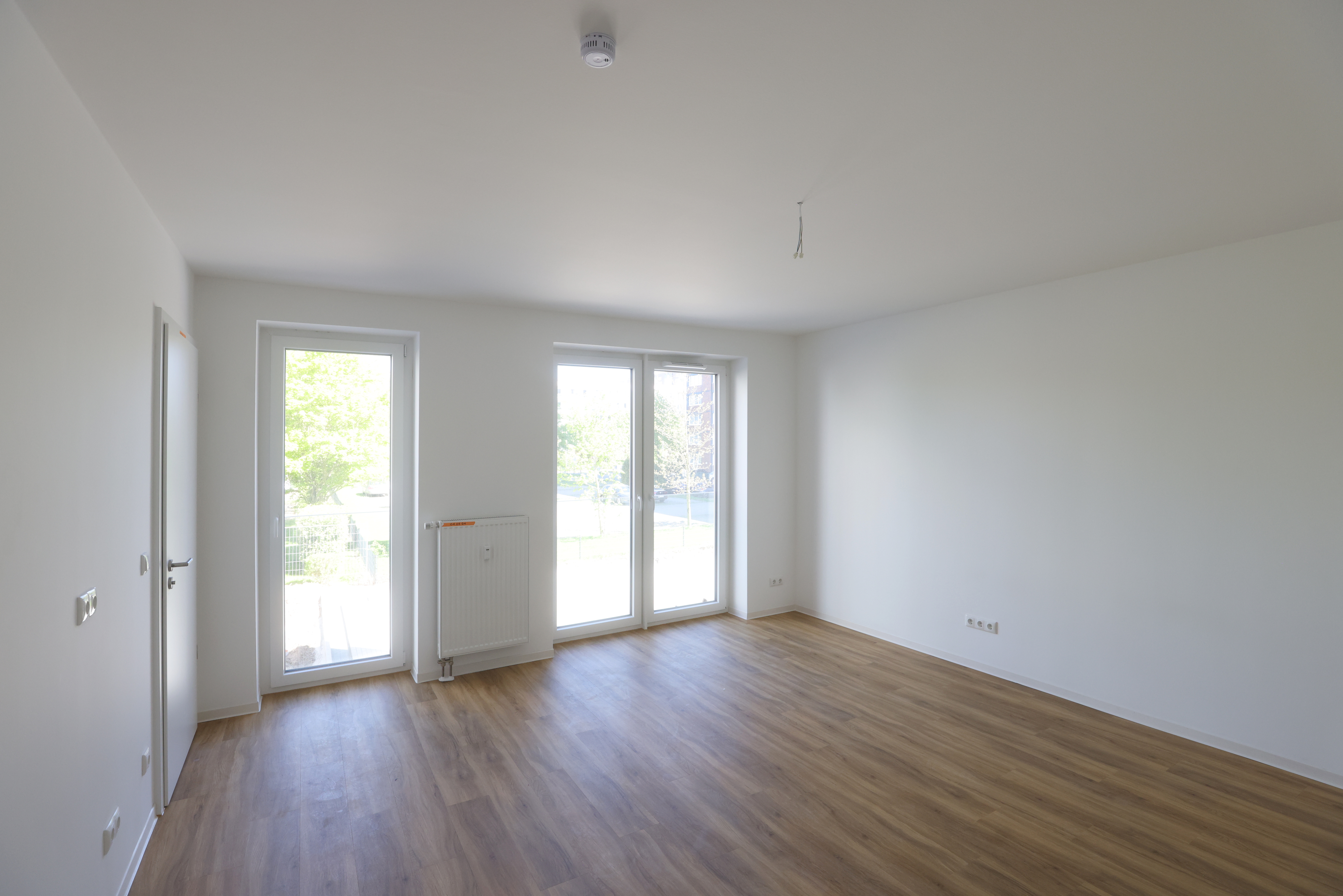 Helles Zimmer mit Fußboden in Holzoptik, viel freier Stellfläche, Zimmertür und drei bodentiefen Fensterelementen.