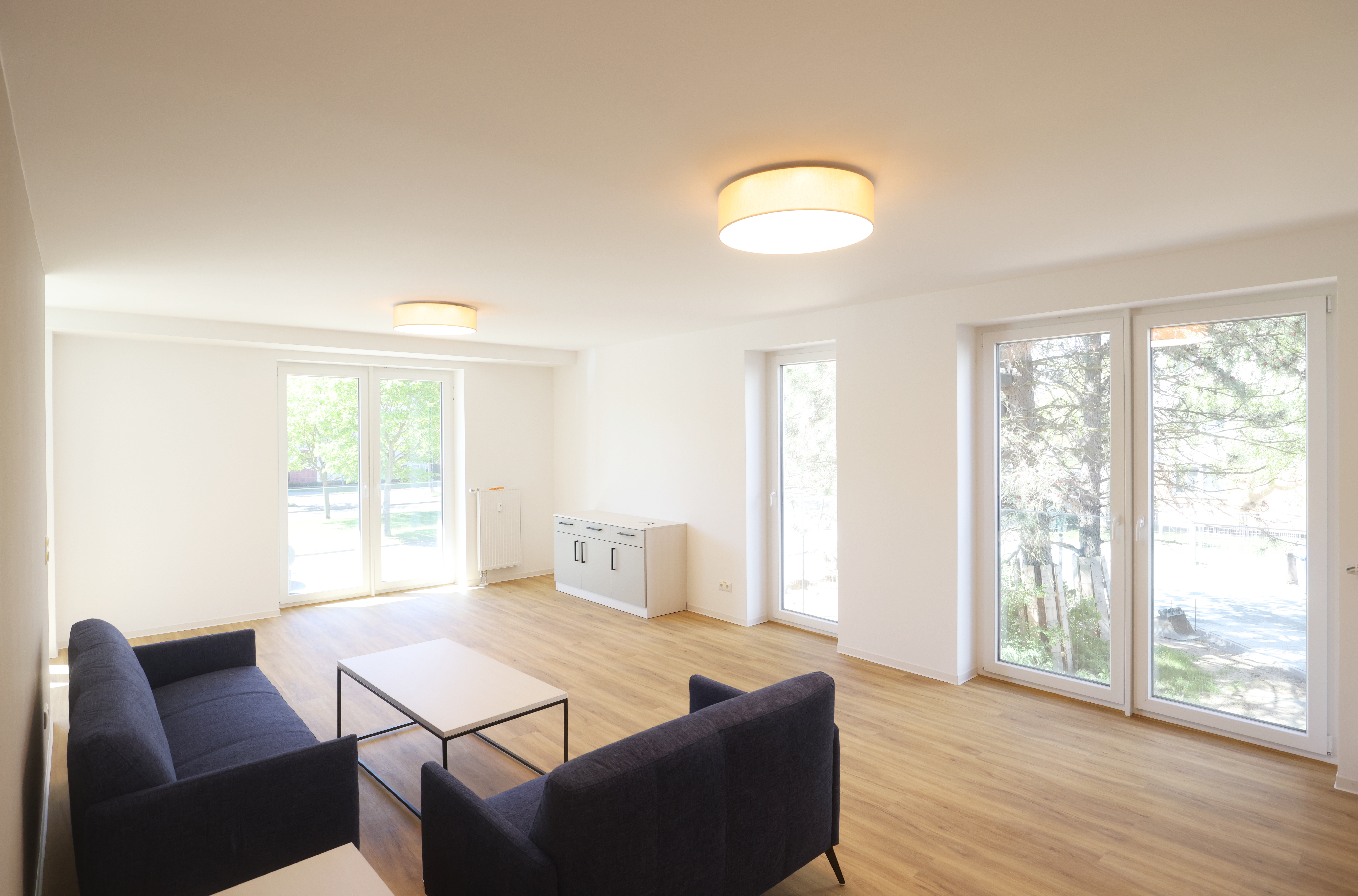 Heller Gemeinschaftsbereich mit Sofas, Couchtisch, Schrank und großen Fenstern bis zum Boden.