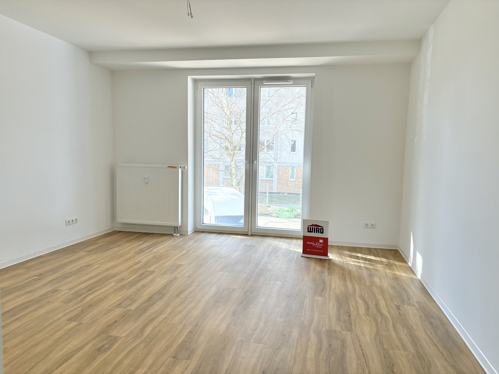 Helles Zimmer mit Fußboden in Holzoptik und bodentiefem Doppel-Fenster. Es sind fünf Steckdosen und ein weißer Heizkörper zu sehen.