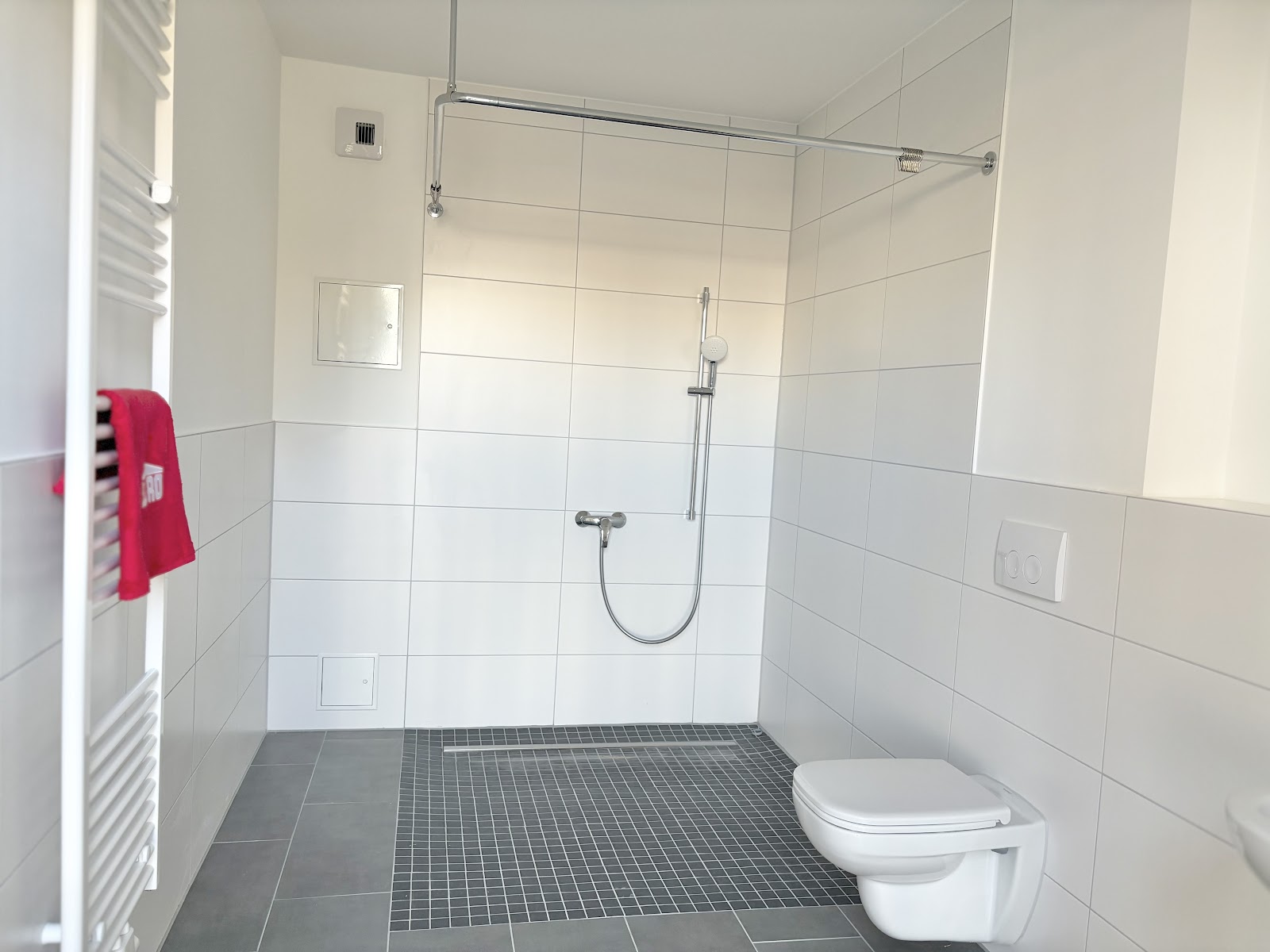Hell gefliestes Bad mit großer bodengleicher Dusche, Duschvorhangschiene, WC, Handtuchheizkörper und freier Bewegungsfläche.