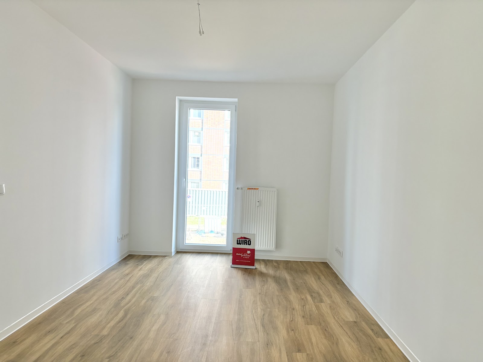 Helles Zimmer mit Holzfußboden, bodentiefem Fenster, Heizkörper und Platz für eigene Einrichtung.