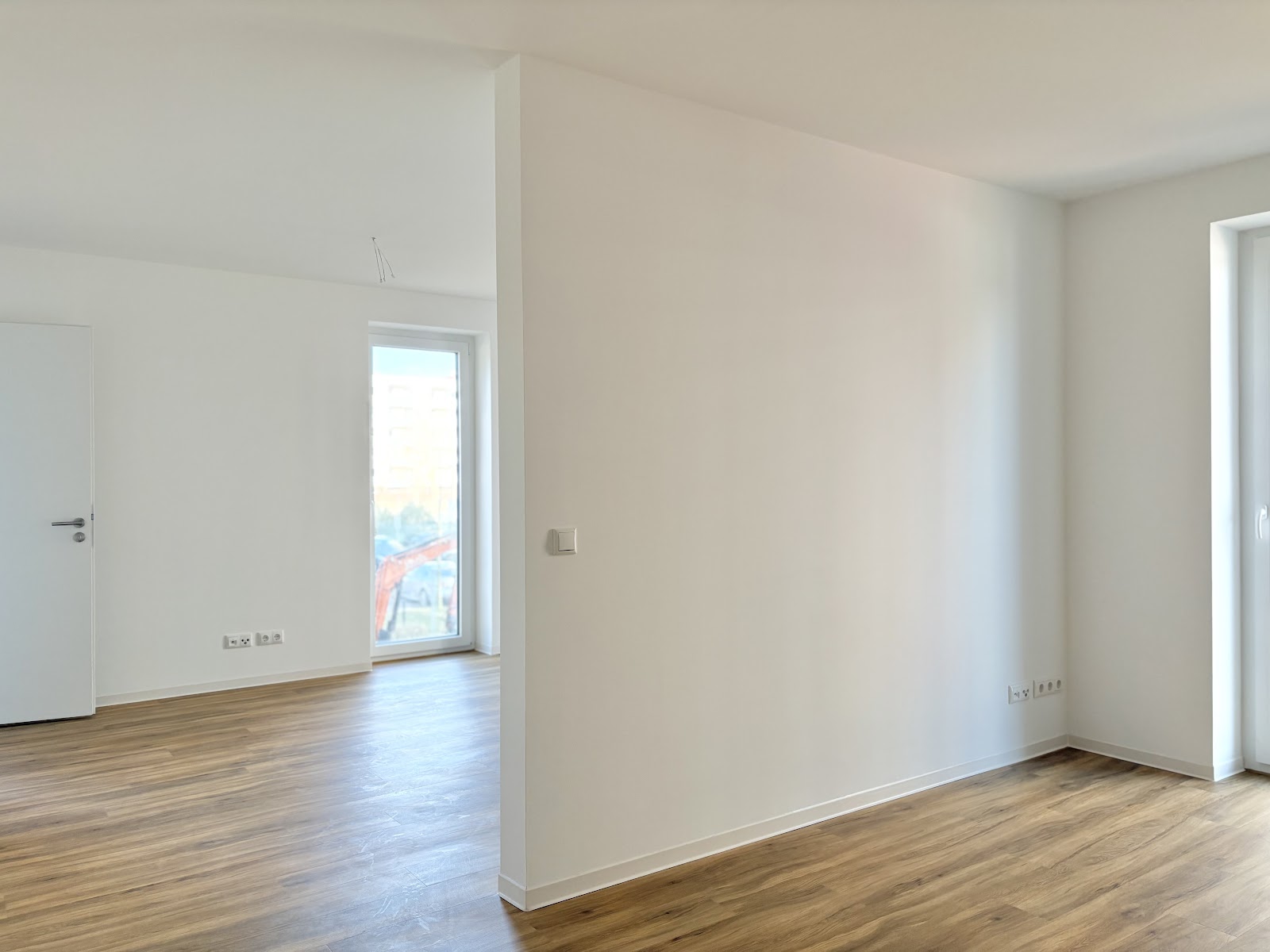 Helles Zimmer mit Holzfußboden, Wandvorsprung zur Raumgliederung, bodentiefen Fenstern und verschiedenen Elektro-Anschlüssen.