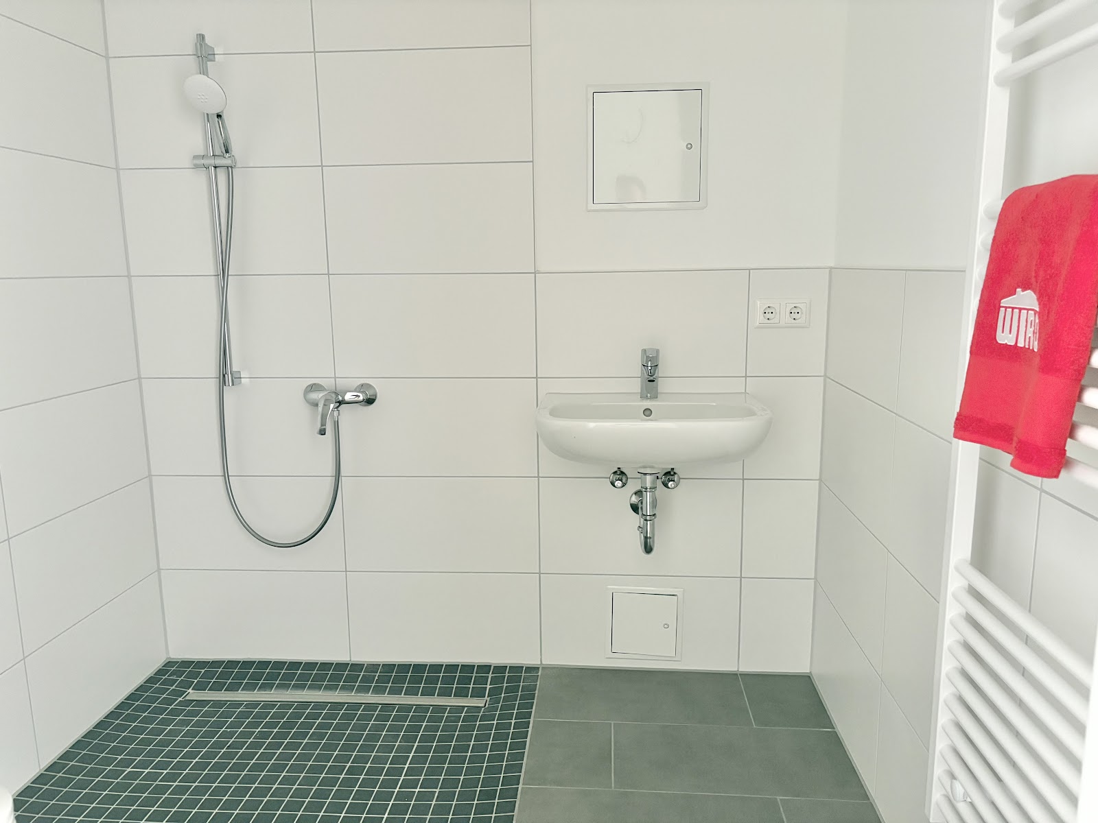 Hell gefliestes Bad mit bodengleicher Dusche, ebener Duschfläche, Waschbecken, Handtuchheizkörper und freier Bewegungsfläche.