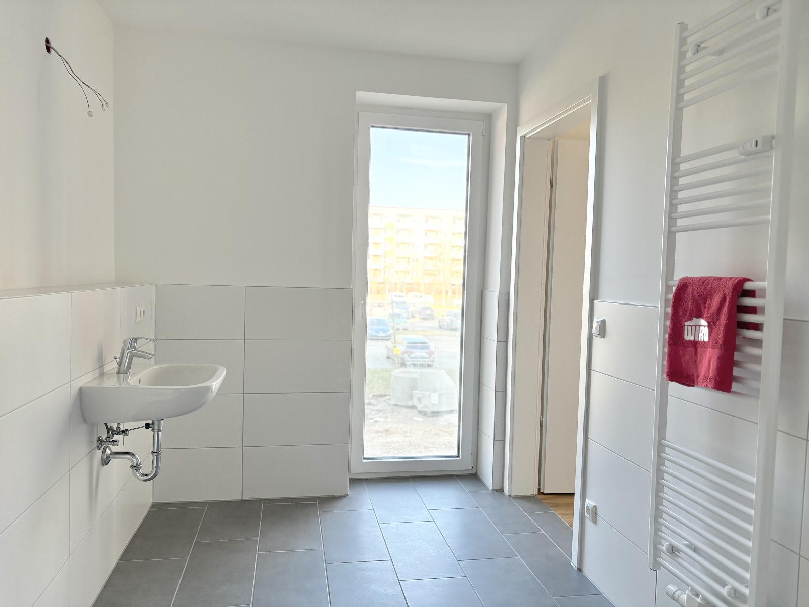 Helles Bad mit Waschbecken, bodentiefem Fenster, Handtuchheizkörper, grauem Fliesenboden und Zugang zum Zimmer.